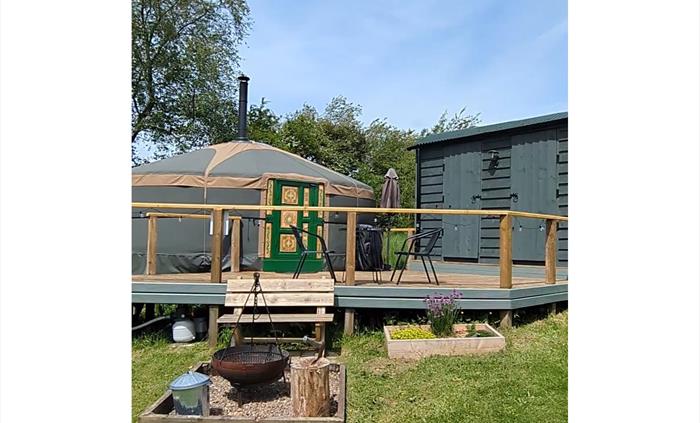 a steel fire pit with tripod in a pebbled enclosure in front of a wood bench, which is in front of a raised deck with a yurt and hut off.